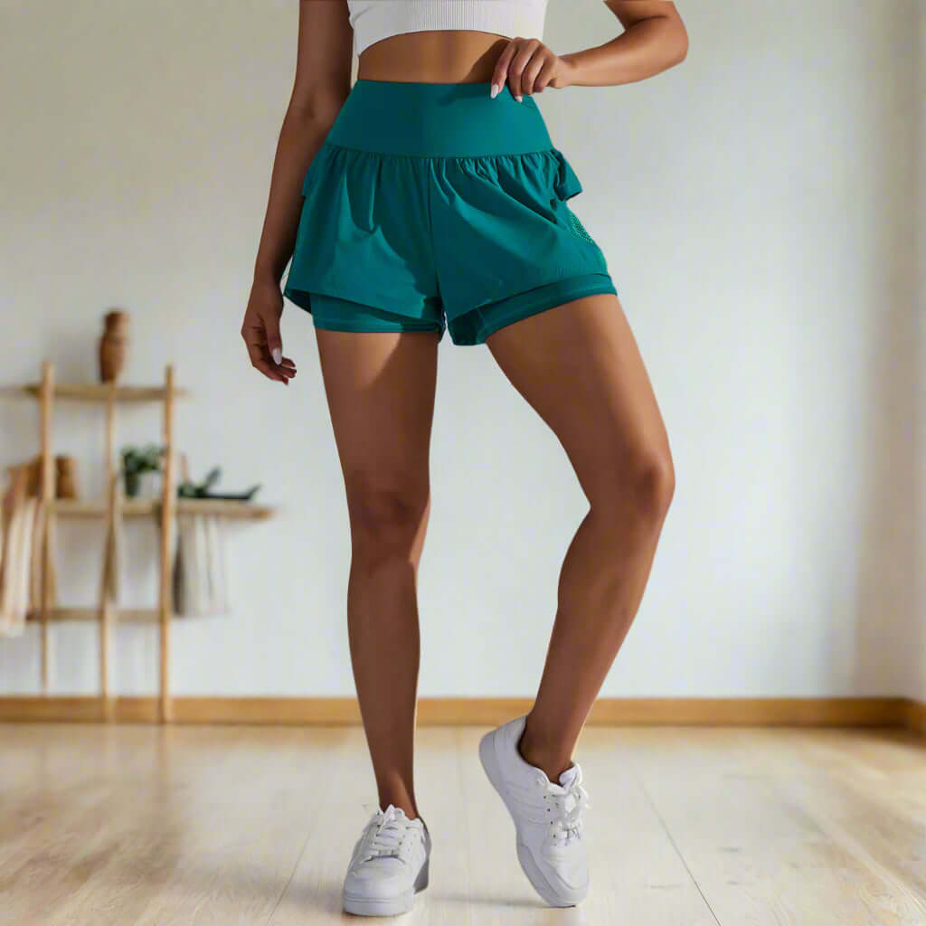 Woman wearing teal double-layer shorts, high-waist design, suitable for yoga or running, showcasing breathable fabric and sleek fit.