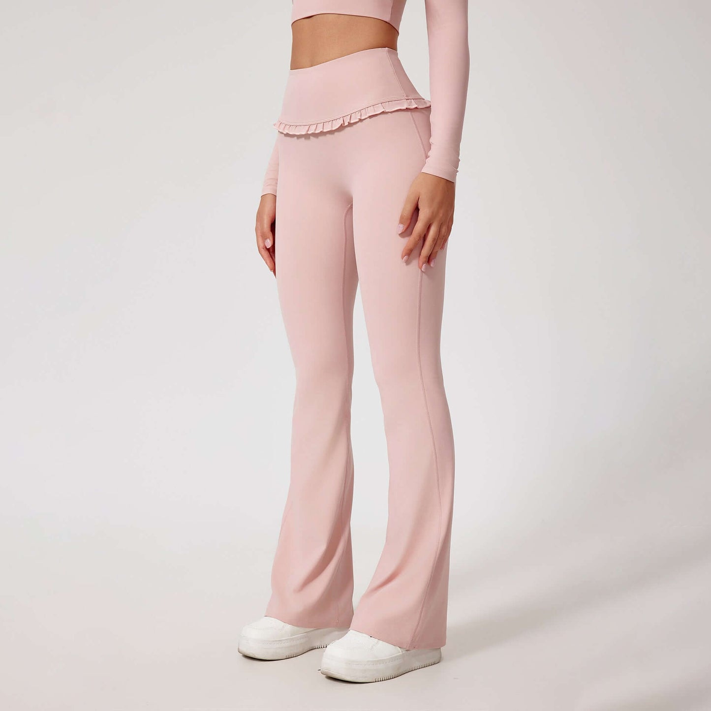 Woman wearing high waist pink leggings with ruffled waistband, paired with matching top and white sneakers, posing on white background.