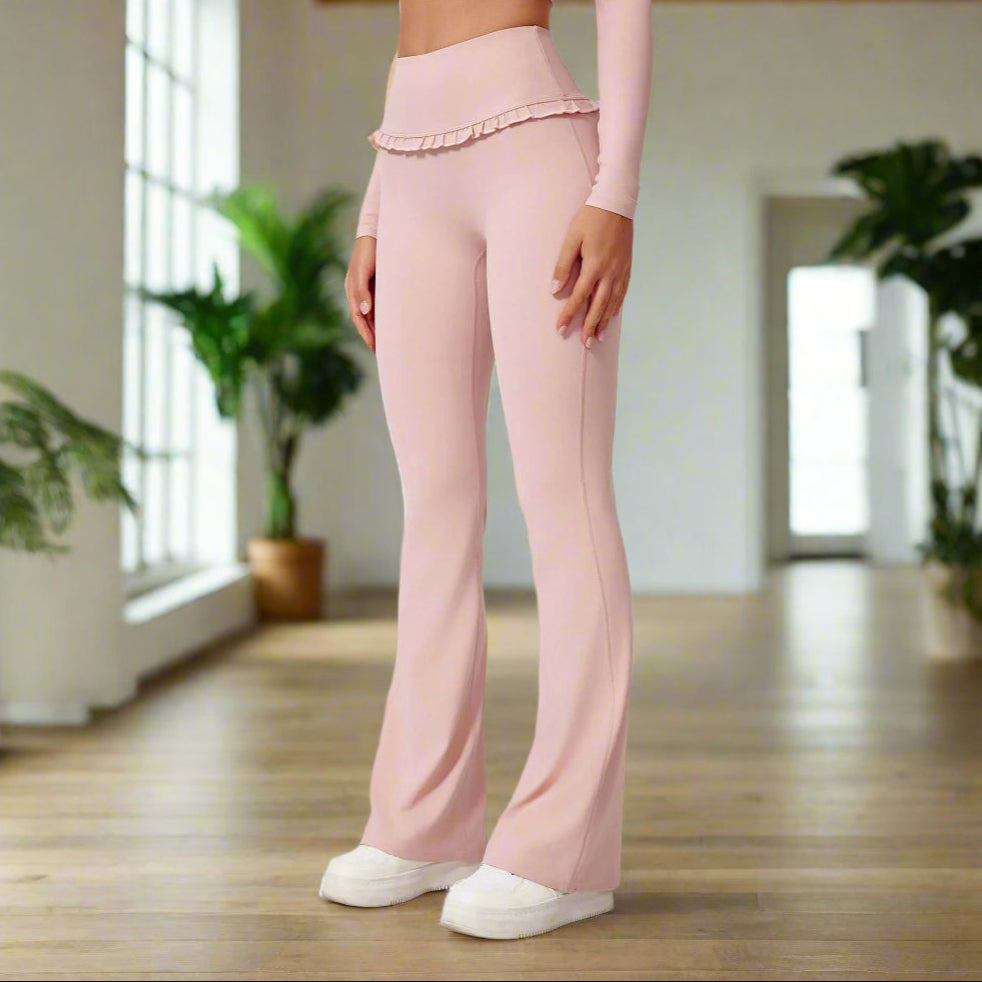 Woman wearing high waist pink leggings with ruffled waistband, paired with matching top and white sneakers, posing on white background.