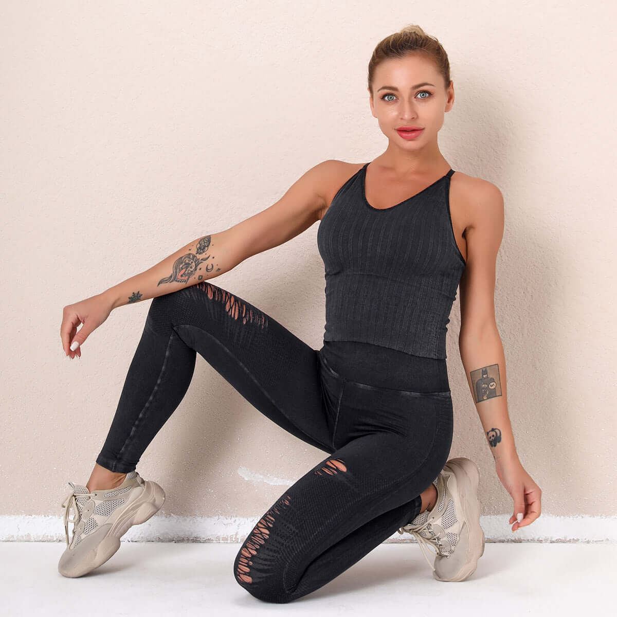 Woman wearing a black seamless suit with ripped details, kneeling and smiling, showcasing activewear fashion against a neutral background.