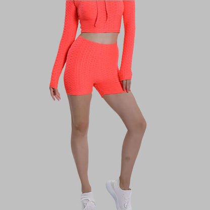 Woman wearing high waist neon orange shorts and matching long-sleeve top with white sneakers on a gray background.