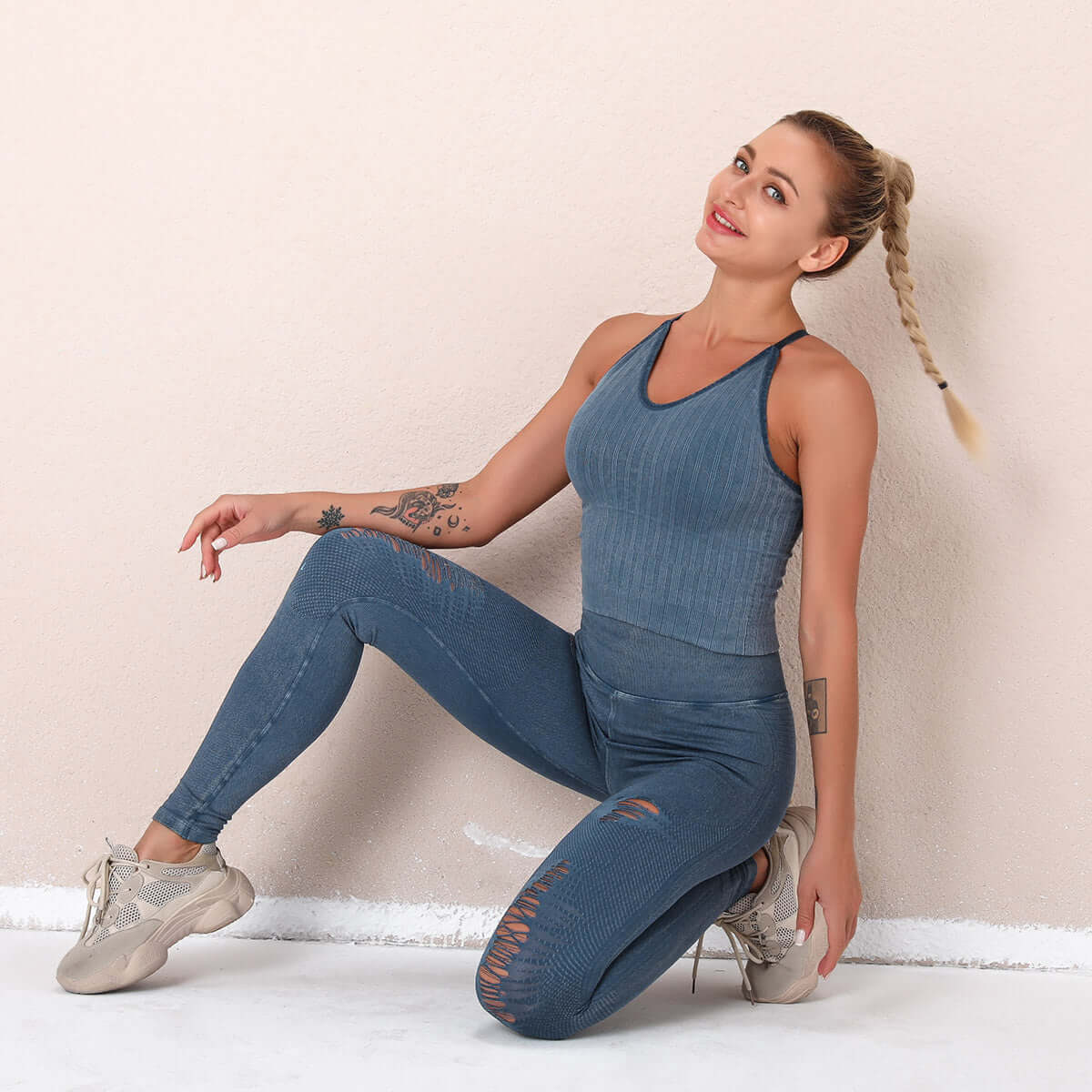 Woman wearing blue seamless fitness suit, kneeling and smiling against a light wall, showcasing athletic wear and style.