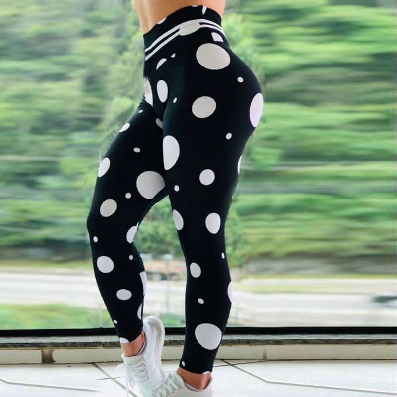 Woman wearing black and white polka dot leggings, standing in front of a window with a blurred green landscape background.