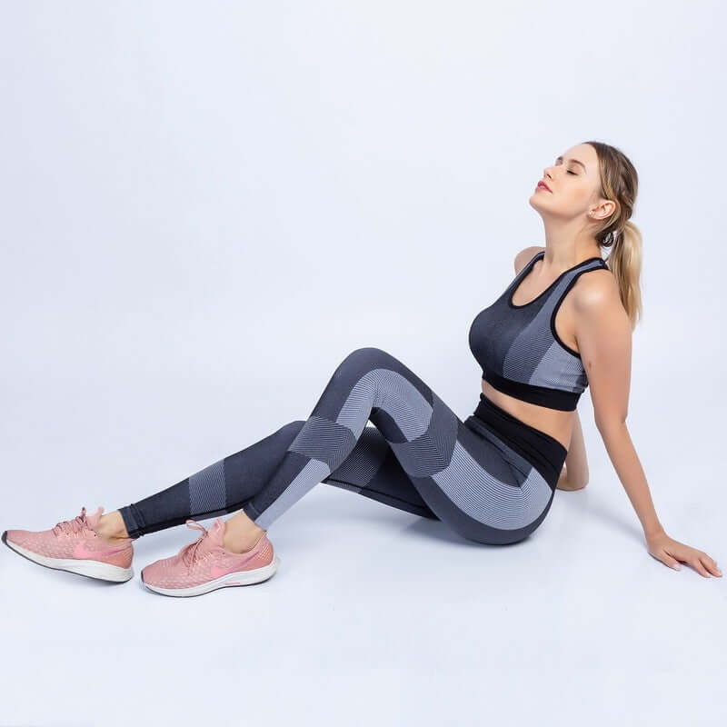 Woman wearing a black and gray seamless yoga set, sitting relaxed with pink sneakers, showcasing sporty and breathable fabric.