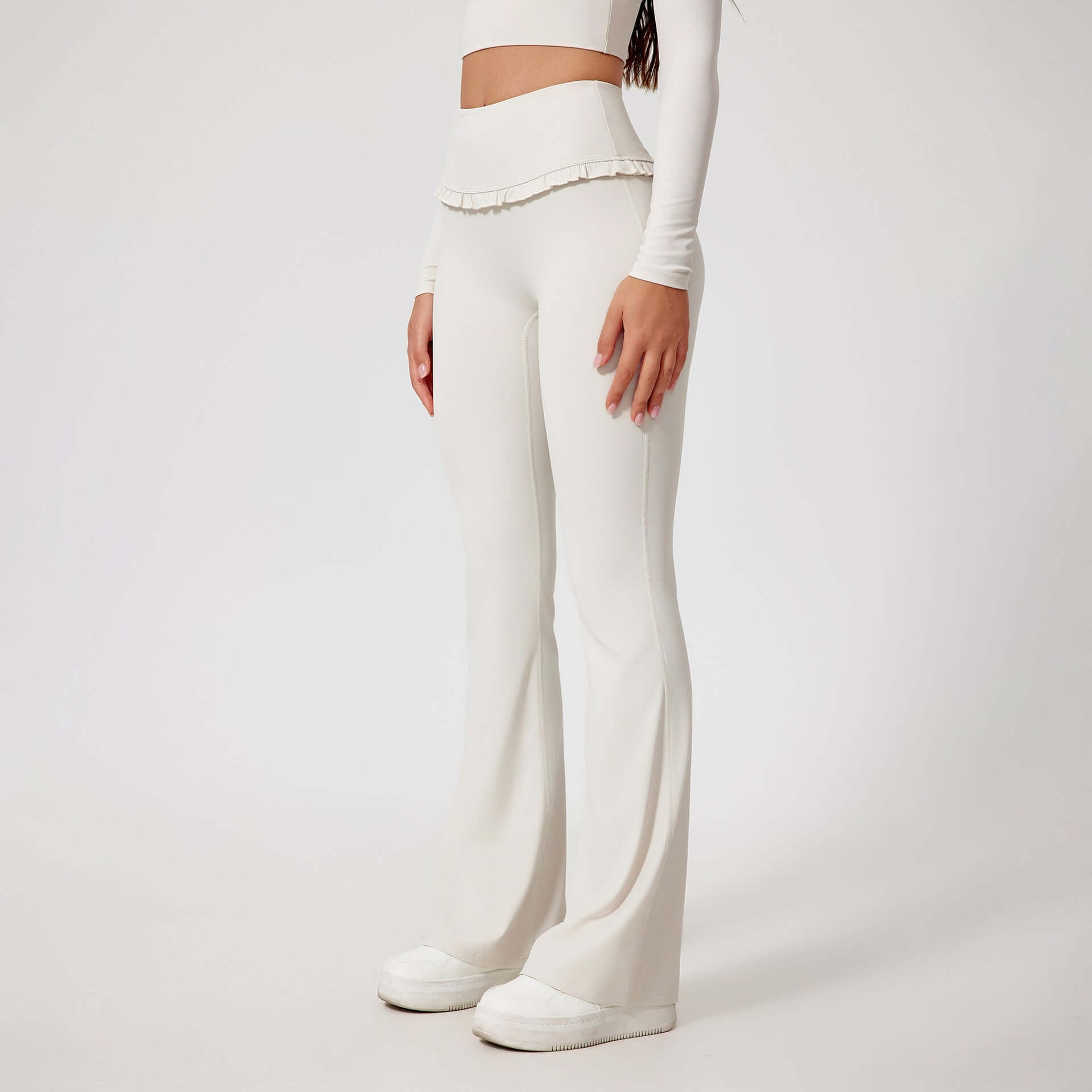 Woman wearing high waist white leggings with ruffled waistband, paired with a long sleeve crop top and white slip-on shoes.