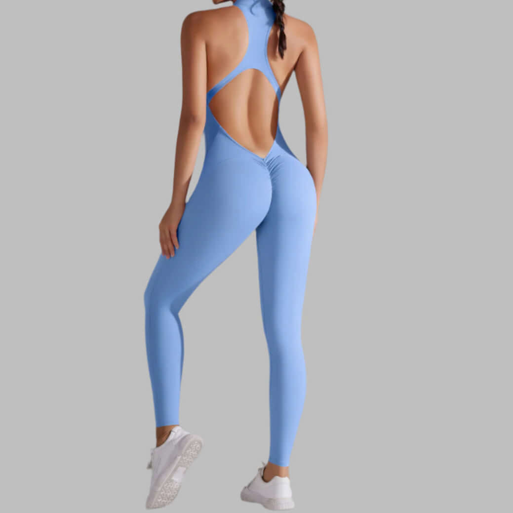 Back view of a woman in a blue seamless workout jumpsuit with open back design, wearing white sneakers against a grey background.